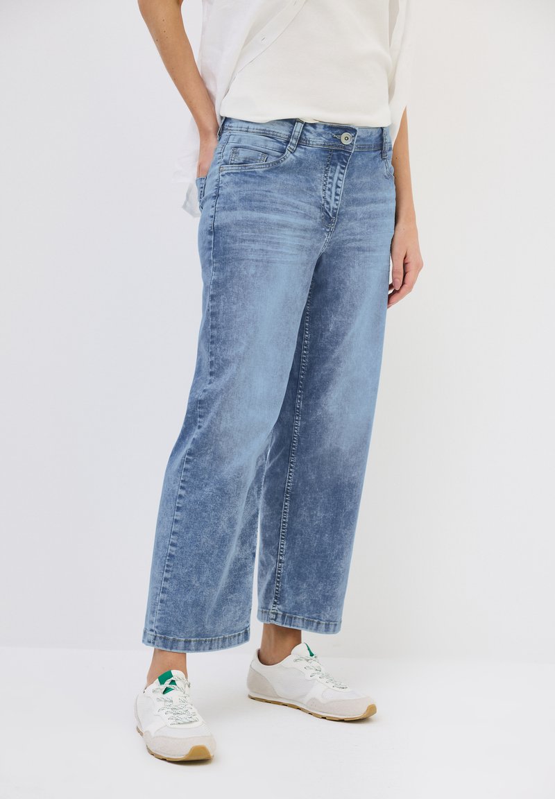Jeans de denim azul claro con un ajuste holgado y longitud recortada. Presenta bolsillos frontales y cierre de botón. Se llevan con zapatillas blancas.