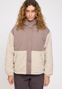 Padded jacket with a brown and cream color scheme, featuring a high collar, zippered pockets, and a soft, textured exterior.