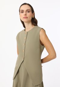 Gilet sans manches de couleur olive, à coupe droite, avec six boutons dorés, une poche latérale et un tissu lisse et texturé.