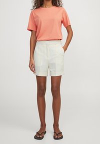 Korallfärgad bomulls-t-shirt i kombination med höga, krämfärgade shorts med uppvikta fållar. Modellen har svarta flip-flops och händerna i fickorna.