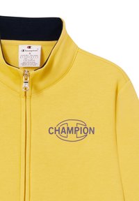 Chaqueta amarilla con cremallera y cuello azul marino, con un destacado logo "CHAMPION" en púrpura. Fabricada con un tejido suave y texturizado.
