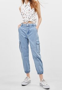Maglietta bianca leggera senza maniche con colletto e nodo frontale. Pantaloni cargo azzurri chiari con polsini elasticizzati, dotati di tasche laterali e vestibilità rilassata.