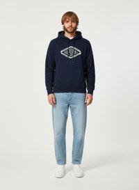 Felpa in cotone blu navy con tasca frontale e lacci, caratterizzata da una grafica del logo bianco. Abbinata a jeans in denim chiaro e sneakers bianche.