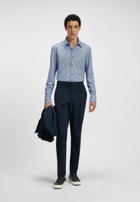 Camicia a righe blu e bianche, pantaloni eleganti blu scuro, tiene un giubbotto scuro, abbinato a sneaker nere, su uno sfondo neutro.