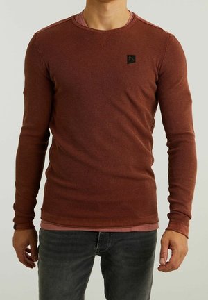 Mężczyzna w rdzawej, teksturowanej koszuli z długim rękawem, na różowej podkoszulce i szarych dżinsach, stojący na jednolitym tle.