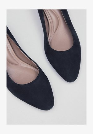 Juodi zomšiniai aukštakulniai su smailiu pirštu ir lygiai tonuotas kremine vidpadžiu. Dizainas pasižymi žemu pjūviu ir švelnia tekstūra.