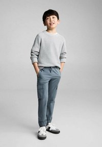 Šedý pletený svetr s manžetovými rukávy, kombinovaný s modrými cargo kalhotami se bočními kapsami. Nosené s černobílými teniskami. Jednoduché pozadí.