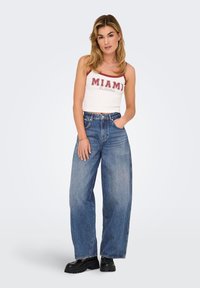 Top corto bianco con la scritta "MIAMI" in lettere rosse, abbinato a jeans di denim blu larghi e stivali neri chunky. Accenti di gioielli semplici.