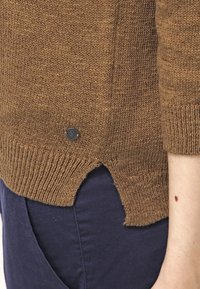 Maglione marrone a maglia con orlo a coste, spacco laterale e un piccolo bottone con logo nero sul bordo inferiore. Presenta un tessuto morbido e texture.