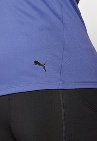 Fialové športové tričko s krátkymi rukávmi, na boku so zobrazeným čiernym logom Puma. Tričko je hladké a priliehajúce, skombinované s čiernymi legínami.