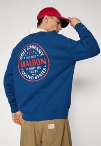 Sweatshirt azul com um logotipo redondo vermelho que apresenta "Golf Company", "Malbon" e "In Golf We Trust". Usado com calças cáqui, mostrando uma etiqueta.