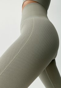 Jasnozielone legginsy z prążkowanej tkaniny mają wysoki stan i teksturowany wzór, podkreślający elastyczność i dopasowanie materiału. Gładkie szwy akcentują design.