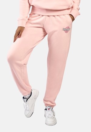 Persoon draagt lichtroze Reebok joggingbroek met elastische boorden en witte Reebok sneakers, staat met één been licht gebogen.