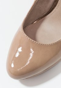 Chaussure à talon haut brillante beige avec un bout arrondi et une surface lisse, présentant un détail subtil de couture le long du bord.