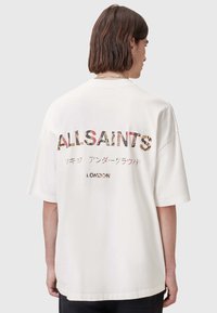 Camiseta blanca de gran tamaño con texto multicolor en la parte posterior que dice "ALLSAINTS", "LONDRES" y otros caracteres adicionales. Textura de algodón suave.