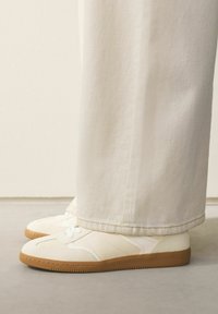 Hvide sneakers med en tekstureret overdel, der har en glat tåhætte og en gummisål i brun. Stylet med bredbukser i kremfarve.