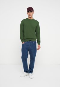 Sötétzöld pulóver bordázott mandzsettákkal, laza kék farmernadrággal párosítva. Fehér sportcipők és bordó baseballsapka teszi teljessé az öltözéket.