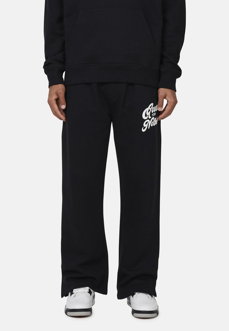 Zwarte sweatshirt met een voorzak, gecombineerd met bijpassende zwarte joggingsbroek met witte tekst aan de linkerkant. Witte sneakers.