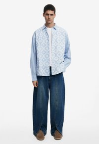 Helles blaues gemustertes Hemd mit Streifen, über einem weißen T-Shirt. Kombiniert mit weiten dunkelblauen Jeans und braunen Slippern.