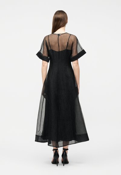 Γυναίκα που φοράει μαύρο διαφανές μεσοφόρι με κοντά μανίκια και μαύρα ψηλοτάκουνα με λουράκι στον αστράγαλο, στέκεται με την πλάτη στραμμένη προς απλό φόντο.