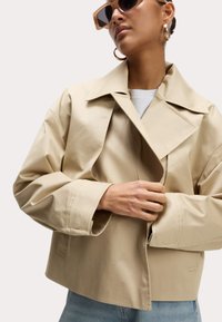 Beige cropped jack met een brede kraag, losse mouwen en een gladde, licht glanzende textuur. Gedetailleerde stiksels en een relaxte pasvorm.