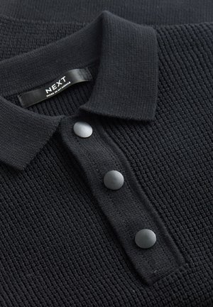 Sort strikket polo med tekstureret stof, tre runde sorte knapper og en krave. Mærket lyder "NEXT, lavet i Myanmar."