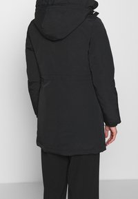 Abrigo acolchado negro con capucha, con una textura suave, ajuste en la cintura y una silueta más larga. Sin detalles visibles de herrajes.