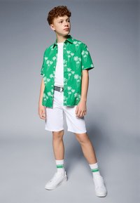 Camicia verde a maniche corte con bottoni e stampa di palme bianche, abbinata a pantaloni corti bianchi e sneakers bianche, accentuata da una cintura nera.