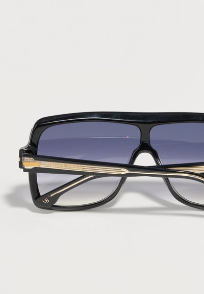 Μαύρα ορθογώνια γυαλιά ηλίου με σκουρόχρωμους φακούς σε διαβάθμιση και χρυσές λεπτομέρειες στα μπράτσα, τοποθετημένα πάνω σε ανοιχτό γκρι επιφάνεια.