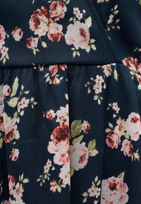 Σκούρο μπλε ύφασμα με ανθωτό μοτίβο που περιλαμβάνει συστάδες από ροζ, κόκκινα και λευκά τριαντάφυλλα με πράσινα φύλλα, με λεπτομέρεια ραφής που δημιουργεί ρυτιδώσεις.