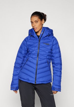 Woman wearing bright blue Columbia Titanium insulated jacket with hood and black outdoor pants, standing against a plain light background.