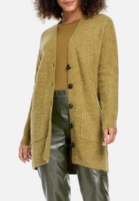 Hořčicově žlutý pletený kardigan s žebrovanou strukturou, předním zapínáním na knoflíky, dvěma bočními kapsami a uvolněným střihem. Spárováno s olivově zelenými kalhotami.