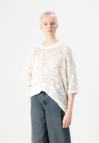 Pull en tricot blanc avec un design géométrique ajouré, coupe décontractée et manches 3/4, associé à un pantalon en denim taille haute.