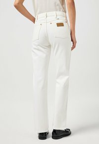 Jeans blancos de talle alto con un parche de cuero en la parte trasera, diseño de cinco bolsillos y pierna recta con una textura suave. Zapatos negros visibles.