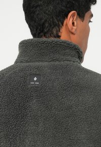 Vīrietis ar īsiem tumšiem matiem, ģērbies tumši pelēkā teksturētā flīsa jakā ar nelielu melnu logotipa uzšuvi augšējā muguras daļā.