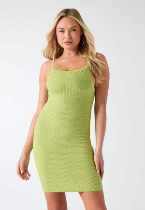 Kobieta z długimi blond włosami, ubrana w limonkową bezrękawnikową dzianinową sukienkę, stojąca z jedną ręką zrelaksowaną, a drugą lekko zgiętą.