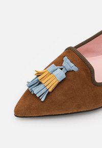Mocassin pointu en daim marron avec des pampilles en daim bleu et jaune et une bordure marron foncé, doté d'une doublure intérieure rose clair.
