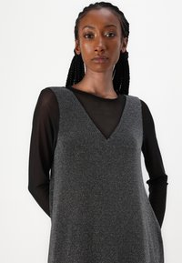 Vestido cinza texturizado com decote em V sobre uma blusa de manga longa transparente preta. O vestido apresenta uma silhueta descontraída e um brilho sutil.