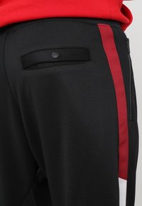 Close-up van zwarte broeken met een verticale rode streep, een knoopzak achterop en een deel van een rode top zichtbaar bij de taille.