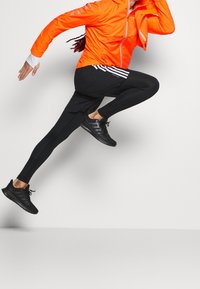 Giacca a vento arancione brillante con dettagli riflettenti, leggings neri aderenti e scarpe da ginnastica nere, che mettono in mostra un movimento dinamico in una posa di salto.