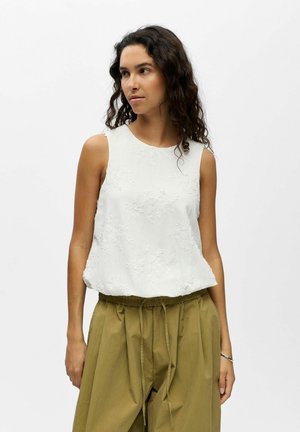 Frau mit lockigem, dunklem Haar trägt ein ärmelloses weißes, floral texturiertes Oberteil und lockere olivgrüne Hosen mit Kordelzug, sie steht vor einem schlichten Hintergrund.