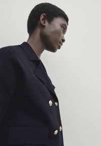 Granatowa marynarka dwurzędowa z złotymi guzikami, z wyraźnymi klapami i teksturowanym materiałem. Model widoczny z boku na jasnym tle.
