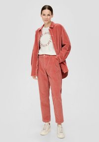 Koszula z koralowej korduroy i dopasowane spodnie, z pionowym ryżem, luźny krój oraz kremowa koszulka z grafiką. Jasnokolorowe trampki.