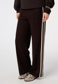 Pantalons large marron en tissu doux, avec des rayures verticales crème et de couleurs mute sur les côtés. Deux poches avant sont visibles.