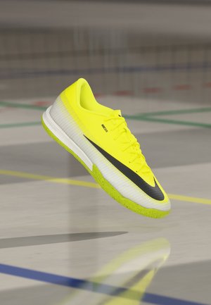 Felgele indoor voetbalschoen met zwarte swoosh en gespikkelde zool, zwevend boven de gymvloer met gekleurde veldlijnen.
