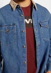 Giubbotto di denim con colletto color beige indossato sopra una maglietta bordeaux con un logo Levi's parzialmente visibile sul petto.