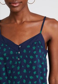 Débardeur sans manches bleu marine avec motif de feuilles vertes, décolleté en V et détail de boutons à l'avant. Fabriqué en tissu léger.