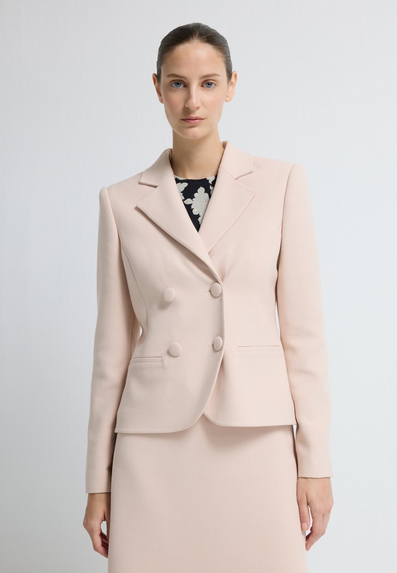 Blazer de doble botonadura en rosa claro con solapas, botones frontales y dos bolsillos con tapa. Tejido suave con un corte ajustado.