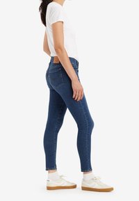 Jeans de mezclilla azul ajustados con cintura alta, con bolsillos traseros y un parche de cuero, combinados con zapatillas blancas y calcetines.