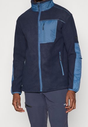 Hombre que lleva una chaqueta polar azul marino con bolsillos en el pecho y en los laterales con cremallera azul claro, y pantalones azul marino a juego con un bolsillo con cremallera.